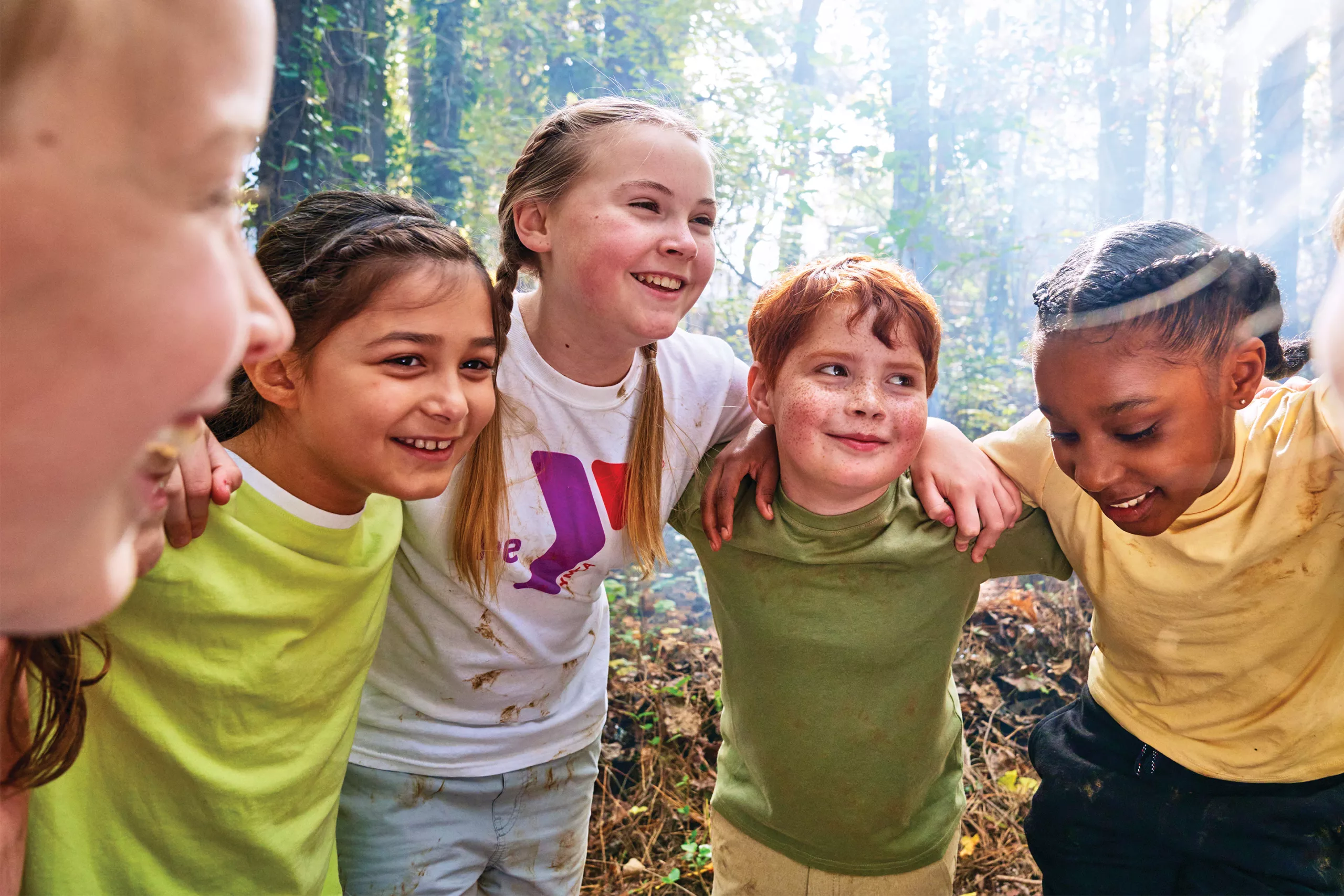 A group of children huddle together with their arms around one another. They are outdoors and smiling.