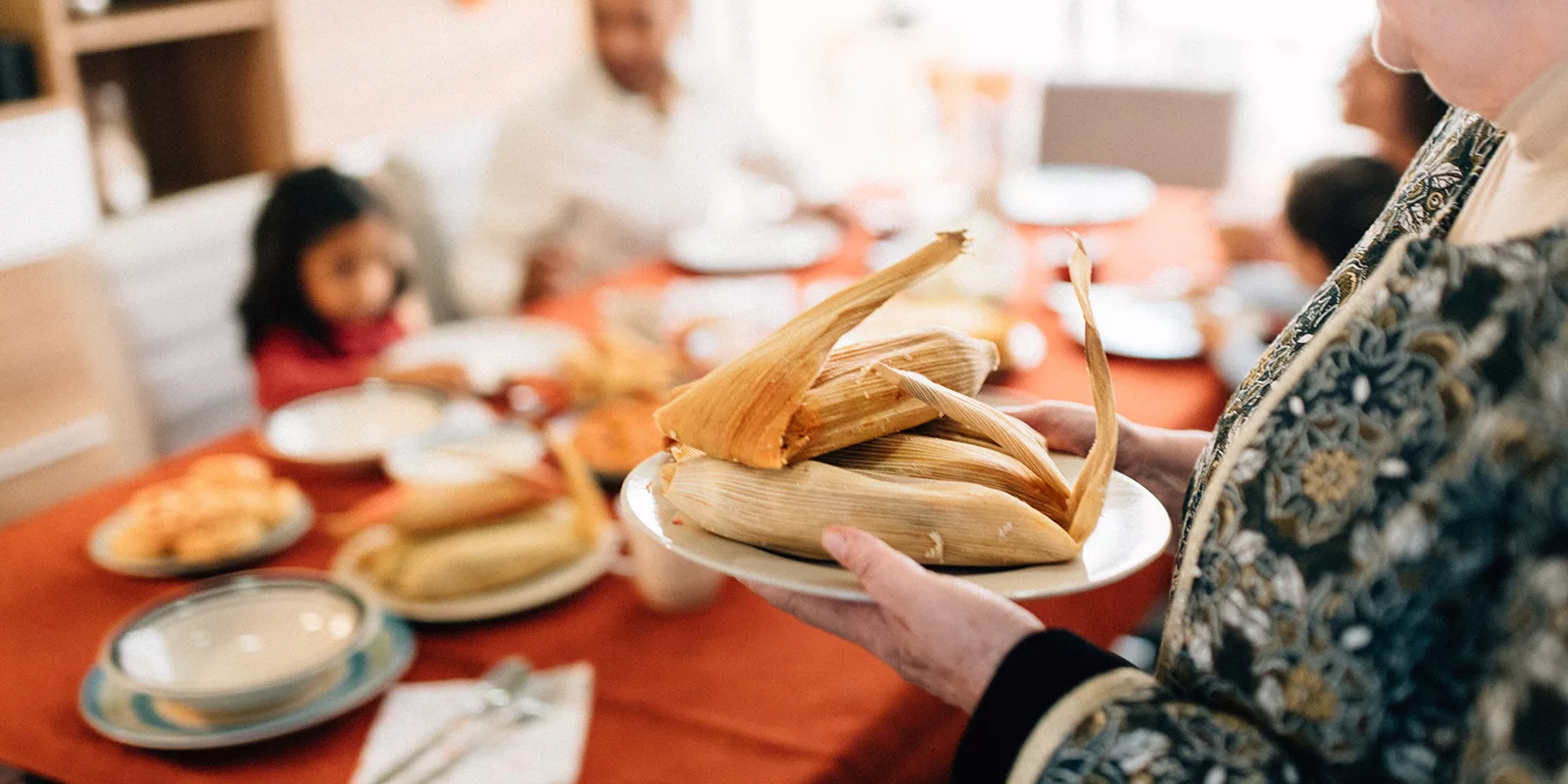 A plate of tamales is seen in focus with a table in the background laid with food and people sitting around it.