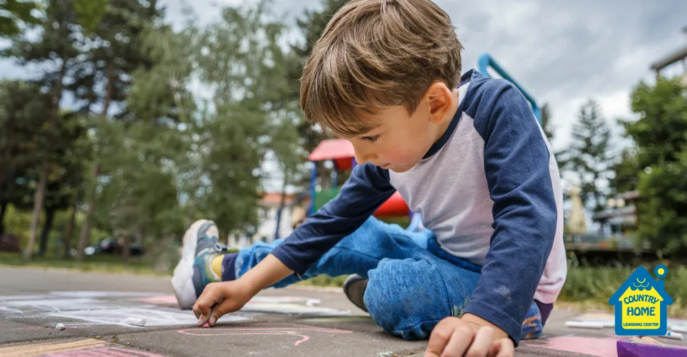 a young boy colors with chalk while sitting on the ground with a park scape behind him