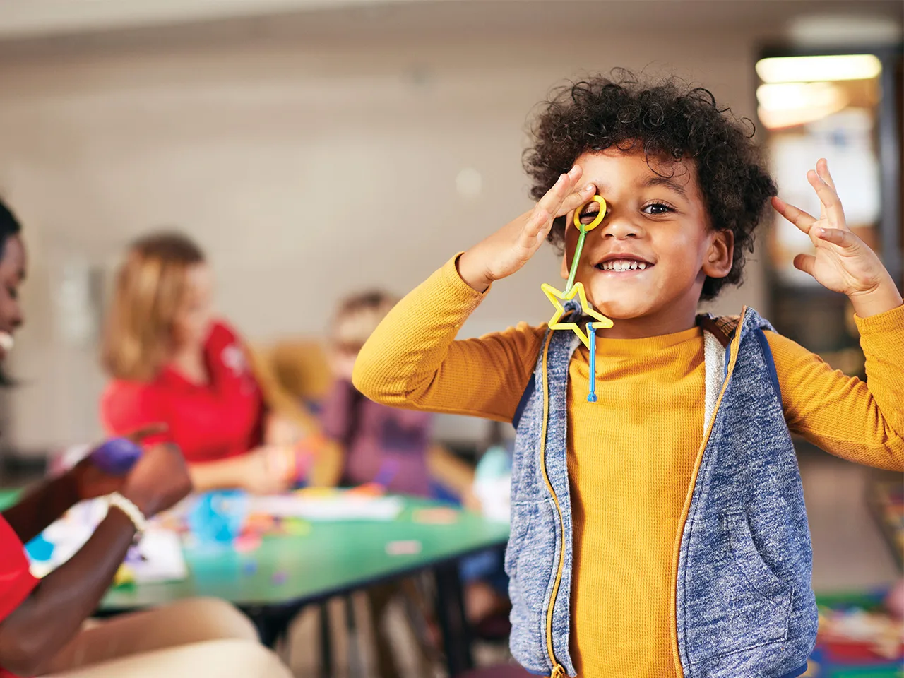 A young child is smiling and playing with toys while Y staff smile and play with other children in the background.