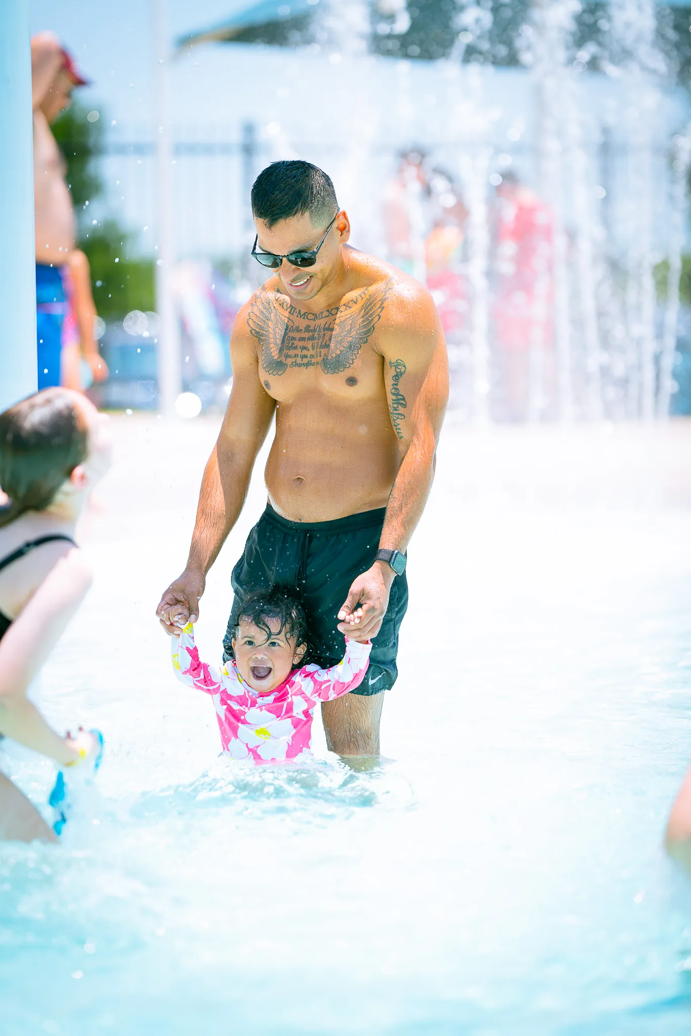 A man stands in an outdoor pool holding his toddler child's hands. The child is smiling with delight.