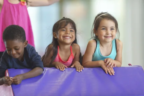 A diverse group of toddlers leans over a soft-play shape inside a gym, smiling happily.